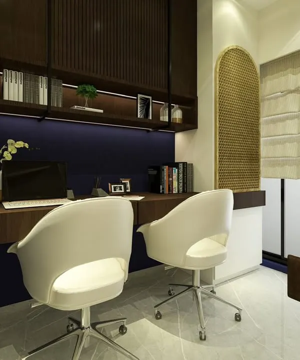 This home office is designed for two, with a long, shared desk against a deep blue wall. Overhead shelving with integrated lighting provides ample storage and a warm ambiance, while the modern white chairs offer comfortable seating.
