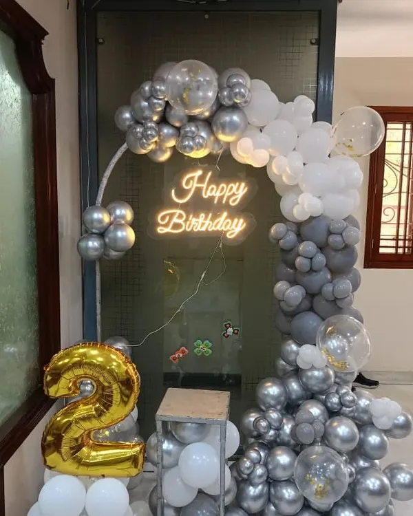A chic and modern birthday setup for a home. The half-arch of grey and white balloons is accented by a "Happy Birthday" neon sign and a large gold foil number '2'.
