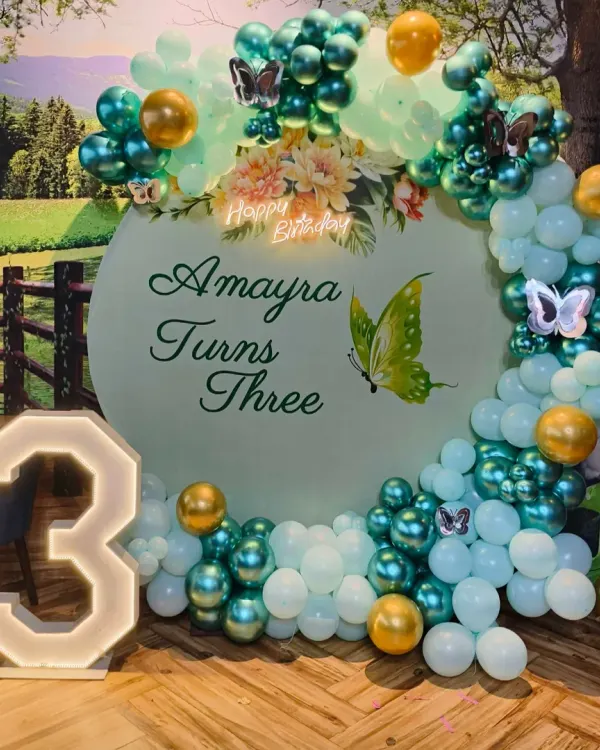 A beautiful butterfly theme for a third birthday. This setup features a custom backdrop, a teal and white organic balloon arch with gold accents, and a large light-up number '3'.