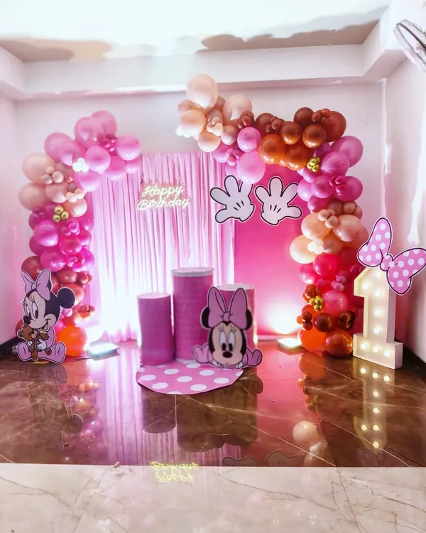 Here is a full view of a Minnie Mouse themed first birthday party. The setup includes a complete balloon arch in shades of pink, a matching pink curtain backdrop, a light-up number '1', and adorable cutouts of Minnie to create a picture-perfect setting.