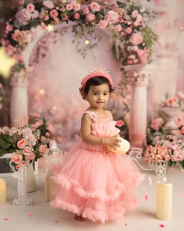 A princess party for a first birthday. This little girl, dressed in a beautiful pink gown, stands in a fairytale setting complete with a floral arch, candles, and a tiny crown.