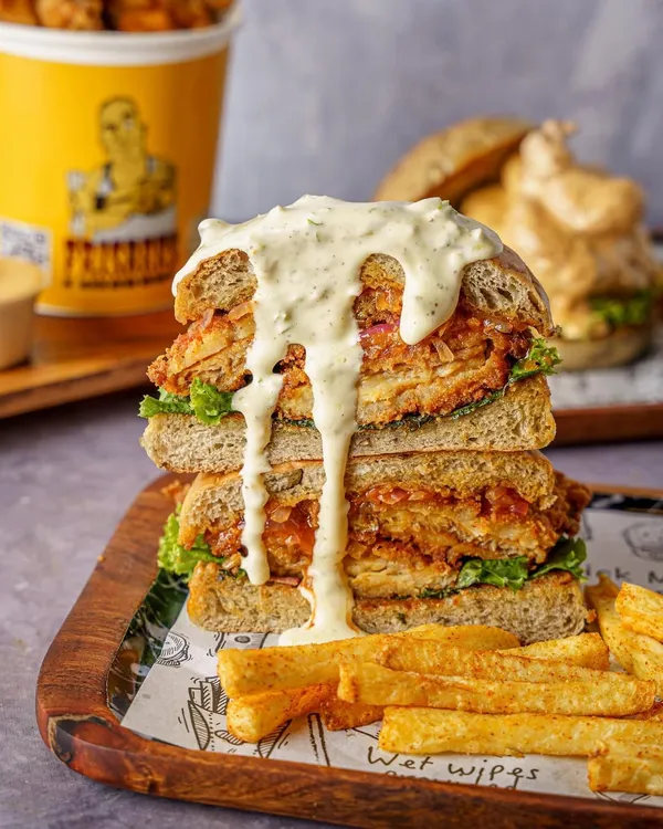 A towering fried chicken sandwich shot for Frisbees Burger Peddler. This image is styled to look indulgent and satisfying, perfect for a new menu launch announcement on social media.