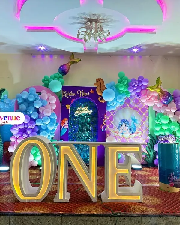 An enchanting mermaid-themed first birthday party. This elaborate setup includes a large "ONE" marquee table, a shimmering sequin backdrop, an under-the-sea balloon arch, and numerous mermaid-themed props and cutouts.