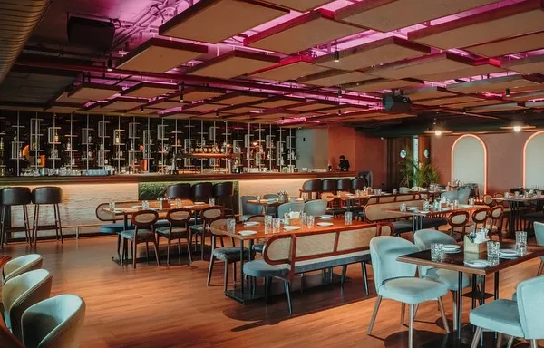 This wide shot captures the dynamic atmosphere of the main dining area and bar. The design uses a mix of seating types, from intimate tables to long benches, all under a ceiling with acoustic panels and dramatic pink lighting.