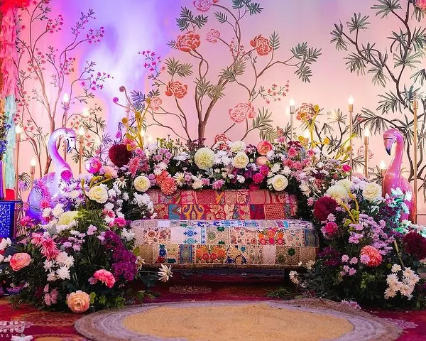 A close-up of a beautiful Mehendi stage with a hand-painted floral backdrop. The decor is enhanced with colorful lighting, a patchwork sofa, and abundant fresh flowers.