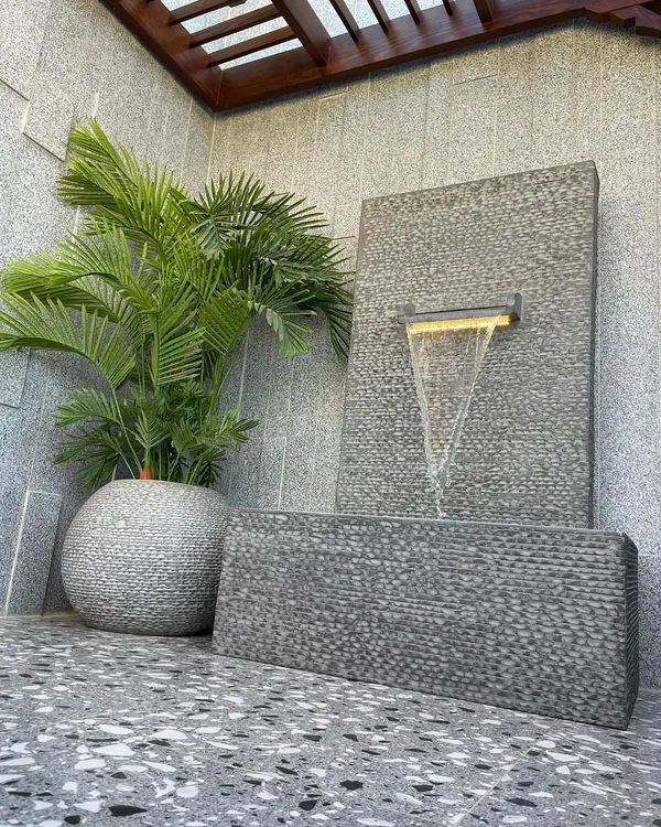 A modern, freestanding water feature made of textured granite. The water cascades gently from a lit spout into a lower basin, creating a peaceful sound and a beautiful focal point, especially at night.