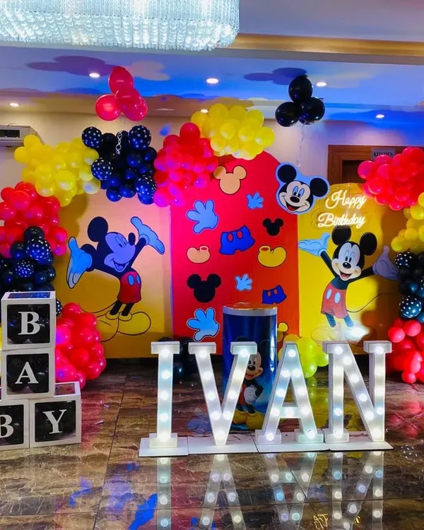 A classic choice for a first birthday, this Mickey Mouse themed party is always a hit. The setup includes a vibrant backdrop with Mickey characters, marquee letters for the name 'IVAN', and themed balloon colors.