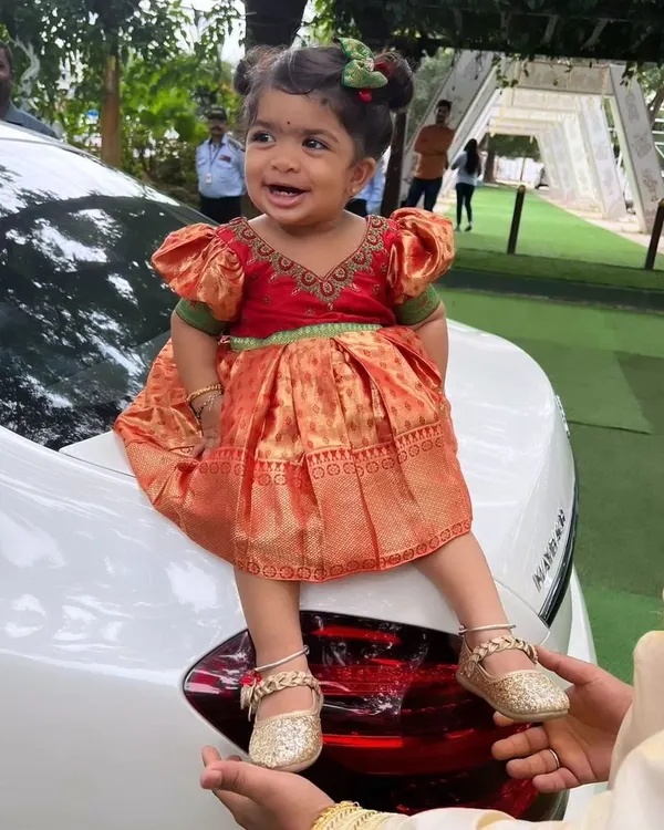 This little princess is all smiles in her custom-designed silk frock. The dress is in a traditional Pattu Pavadai style with a bright red and orange combination and cute puff sleeves.