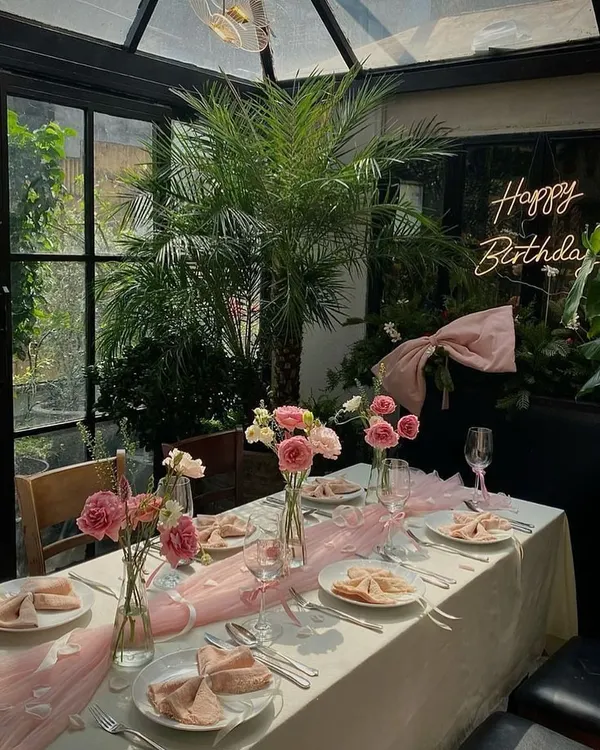 A beautiful birthday dinner setup in a sunroom. We used a soft pink color palette with a tulle table runner, fresh pink roses and carnations, and a "Happy Birthday" neon sign for a chic and celebratory feel.
