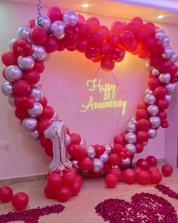 Apni pehli anniversary ko khaas banane ke liye humne yeh red aur silver chrome balloons ka heart shape frame taiyar kiya hai. Beech mein 'Happy Anniversary' ka neon sign iski sundarta aur badha raha hai.