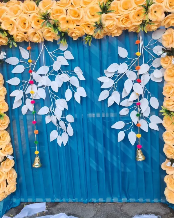 A modern and vibrant pooja backdrop featuring a frame of yellow artificial roses against a blue curtain. The hanging white leaf elements and small bells add a creative touch to the festive decoration.