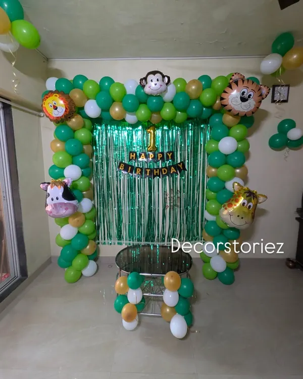 A classic jungle theme birthday decoration for a home setting. This setup includes a green and gold balloon arch, a shimmer curtain backdrop, and cute animal head foil balloons like a lion, tiger, and monkey.