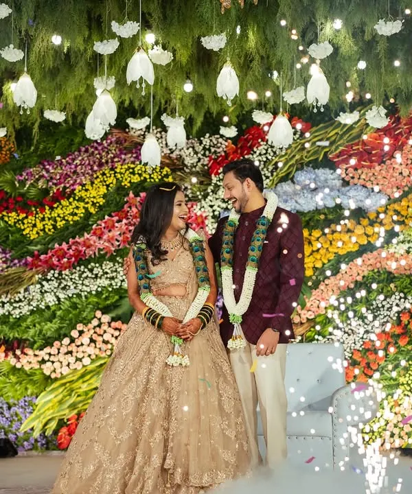 A stunning shot of a couple on their reception stage, set against a massive, vibrant wall of fresh, multi-colored flowers. The falling confetti adds a touch of magic to the moment.