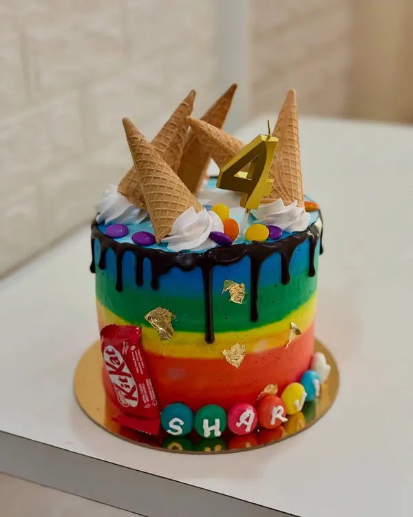 A vibrant rainbow cake that's perfect for any kid's birthday party. This one is topped with ice cream cones, a chocolate drip, and colorful candies for a fun, over-the-top look.