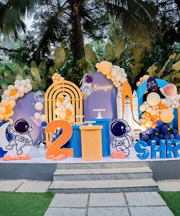 A complete outdoor stage setup for a space-themed second birthday. It features astronaut cutouts, a rocket ship, a telescope, and a vibrant orange and blue color scheme.