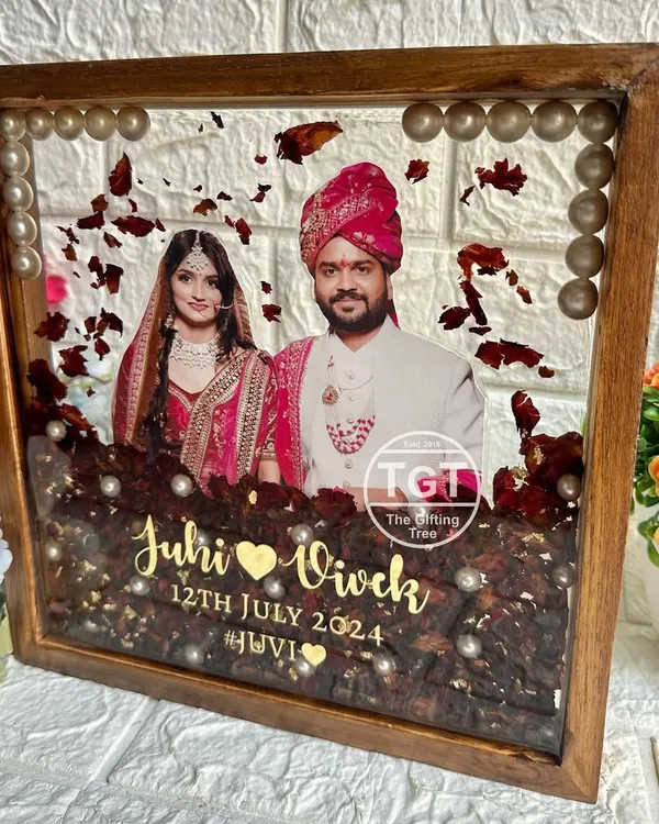 A stunning 10-inch square teakwood frame preserving a couple's varmala. The deep red flowers and pearls are set in clear resin, with a central photo and golden text for the names and date.