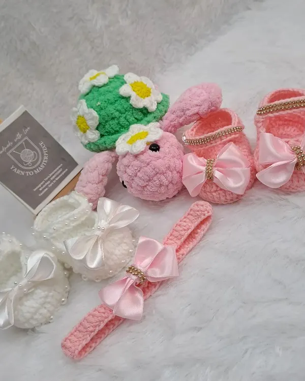 This baby gift hamper is filled with handmade treasures. It includes two pairs of booties with satin bows, one in white and one in pink, a matching pink headband, and a cute turtle toy with a daisy shell.