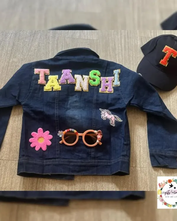 A custom denim jacket for a child named Taanshi. The back is decorated with her name in colorful patchwork letters, along with a unicorn and flower patch. The set also includes a matching initial cap.