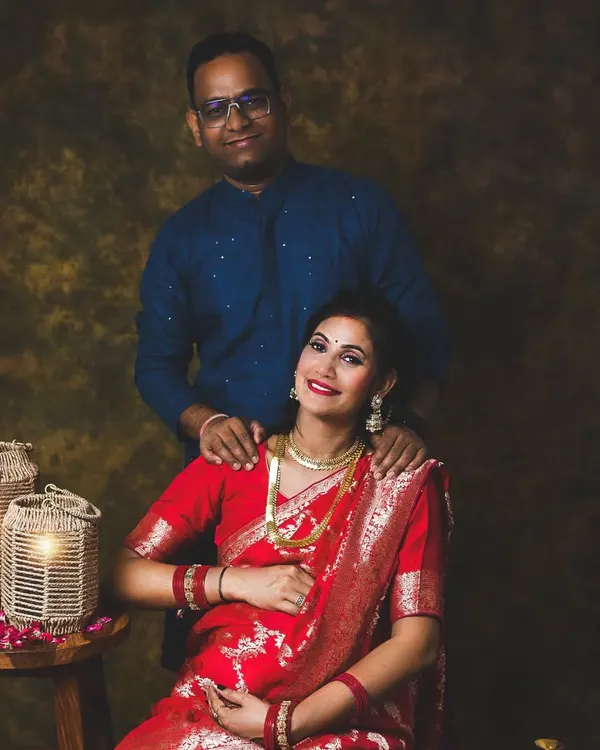 A beautiful traditional portrait of an expecting couple. The rich red Banarasi saree looks stunning against the classic painted backdrop.