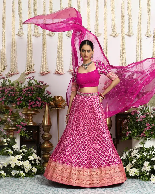 This vibrant fuchsia Bandhani lehenga, with its flowing dupatta and intricate Banarasi border, embodies the joyful spirit of celebration. It is a perfect statement piece for a sangeet or wedding reception, designed for the woman who embraces both tradition and bold self-expression.