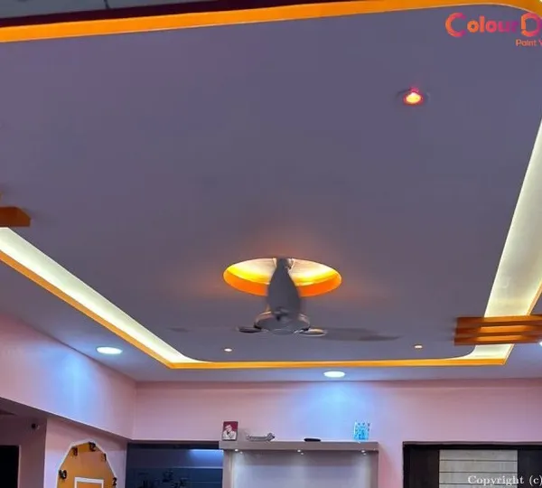 This living room features a multi-level false ceiling with integrated warm and cool lighting. The design incorporates recessed spotlights and hidden cove lighting around the perimeter, creating a bright yet inviting atmosphere while seamlessly accommodating the central ceiling fan.