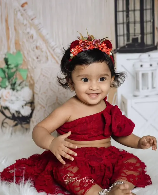 A perfect first birthday portrait! This little girl is all smiles in her red lace outfit, sitting on a fluffy white rug with a bohemian-style backdrop.