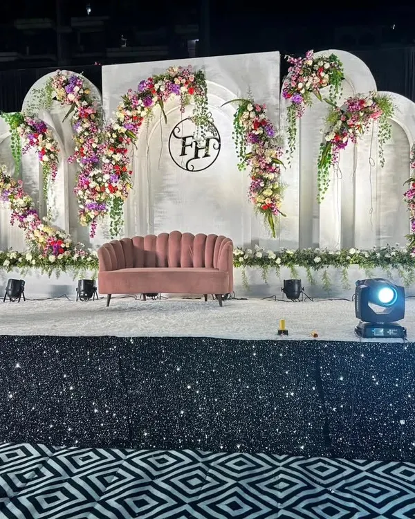 A grand and elegant reception stage we designed, featuring a pristine white backdrop with classic architectural arches. The look is softened with cascading floral arrangements in purple, red, and white, and a custom monogram for the couple, creating a personalized and sophisticated focal point.