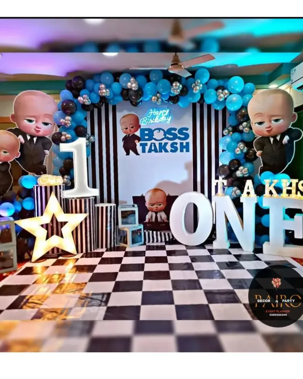 A complete Boss Baby theme for Taksh's first birthday. The setup includes a custom-designed stage with a striped backdrop, checkered flooring, light-up marquee letters, and multiple character cutouts for a fun, immersive photo zone.