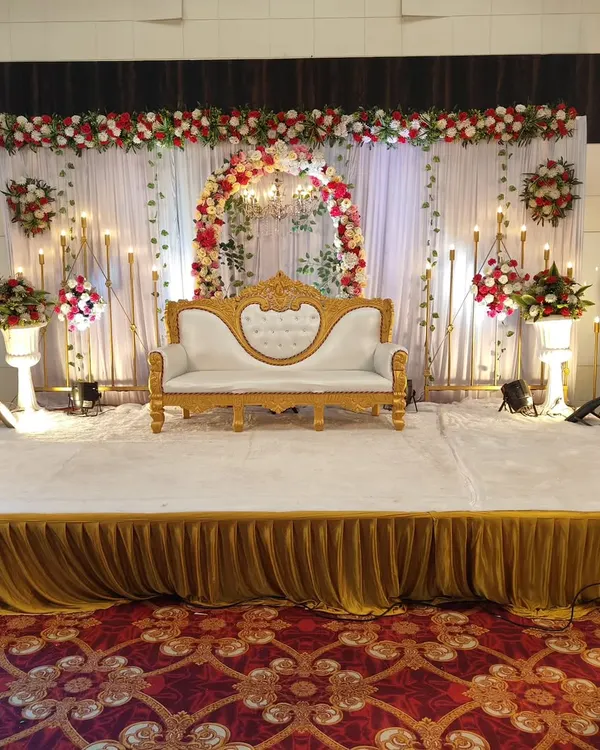 A classic and elegant stage setup for a wedding reception or engagement. It features a beautiful floral arch, a central chandelier, and warm candle-style lighting, all centered around a formal white and gold sofa for the couple.