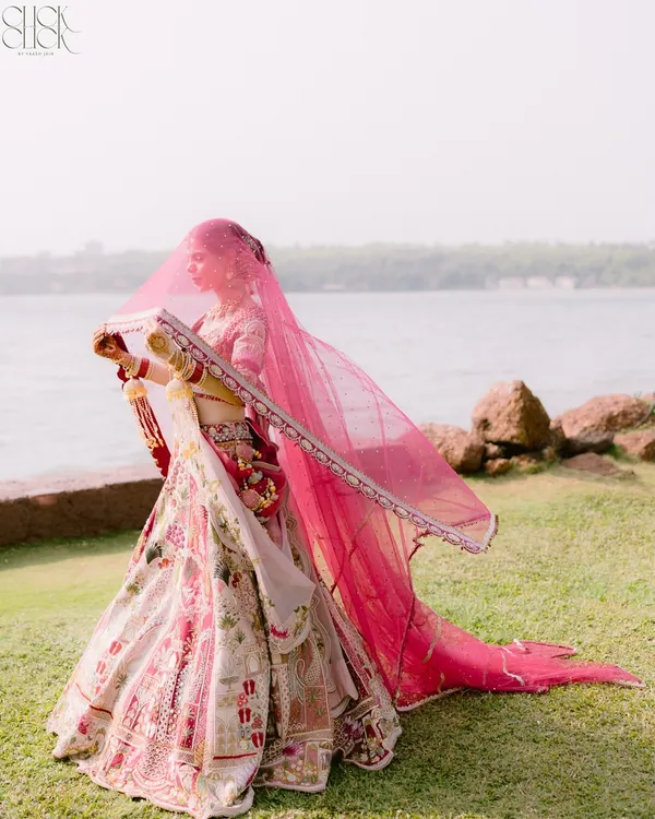 A stunning bridal portrait by the sea in Goa. The bride's pink veil flowing in the wind against the water creates a breathtaking, ethereal image that is both grand and graceful.