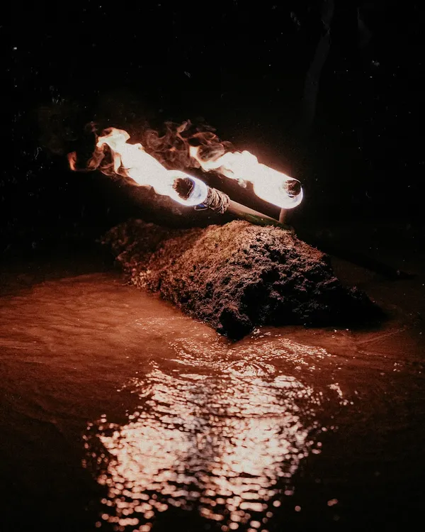 An atmospheric shot of two lit torches resting on a rock in the water at night, their flames reflected in the dark water. This sets the scene for a dramatic and fiery beach pre-wedding photoshoot.