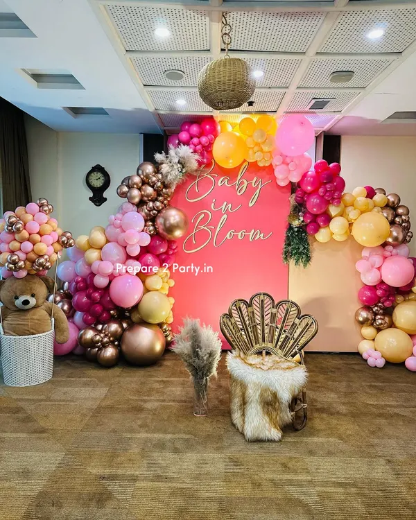 A vibrant and stylish "Baby in Bloom" themed baby shower. The setup features a bold pink backdrop, a stunning balloon arch in shades of pink, orange, and rose gold, and a chic peacock chair for the mother-to-be.