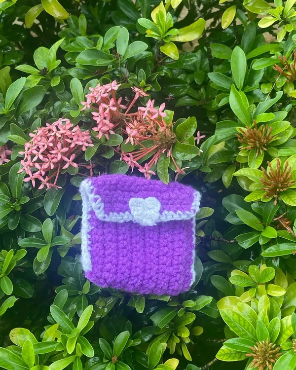 My handmade crochet pouch in purple and white, sitting in a bush. It's perfect for keeping your AirPods, coins, or other small treasures safe.