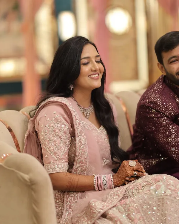 A candid shot of a happy bride at her engagement. Her makeup features a minimal, dewy finish with soft pink tones to complement her pastel pink lehenga, paired with simple wavy hair for an effortless look.