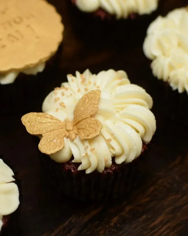A close-up of a red velvet cupcake from the white and gold set. The design features a delicate, handcrafted golden butterfly resting on a swirl of cream cheese frosting, finished with a sprinkle of edible gold dust.