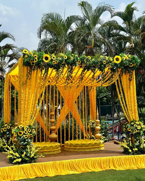 A picture-perfect Haldi mandap. The structure is draped in bright yellow fabric and adorned with strings of marigolds and sunflowers, creating a cheerful and traditional look.