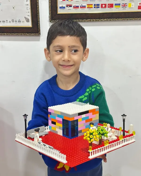 A young boy proudly holds up his impressive LEGO creation: a modern house complete with a detailed garden, fence, and a red carpet entrance. This showcases the architectural and design skills kids can develop.
