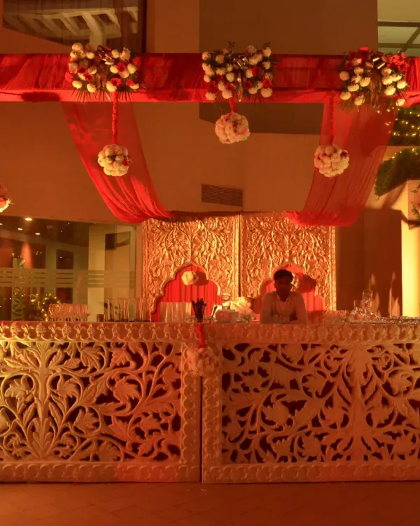 A beautifully crafted bar counter for a traditional wedding. The bar front features intricate white carving, illuminated from within, and is set against a backdrop of ornate golden panels and red drapes.