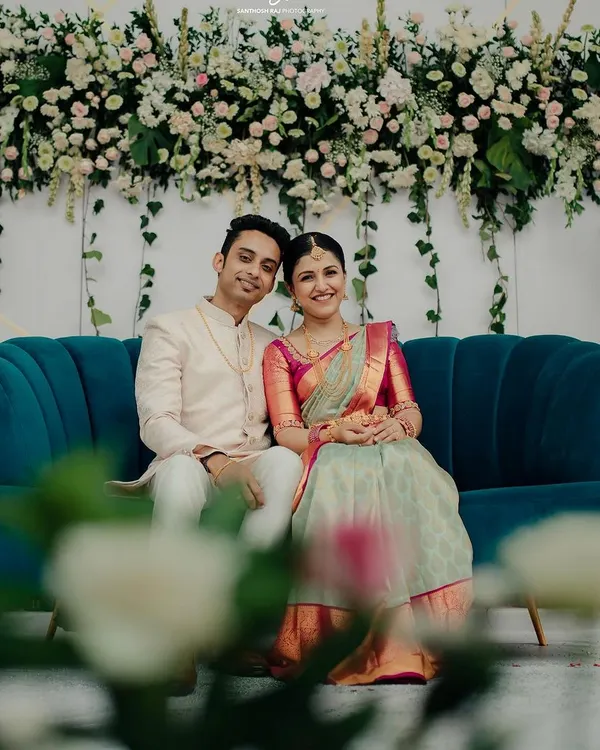 A beautiful moment from Sonika and Abhishek's engagement. The couple is seated on a stylish teal sofa against our custom floral wall, a design that combines white and pink blooms with fresh greenery for a soft, romantic feel.