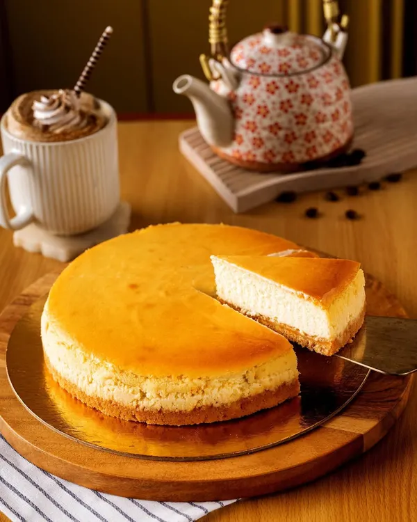 A classic New York cheesecake, beautifully captured for Bisque bakery. The focus is on the creamy texture and golden-brown top, presented simply on a wooden board with a cup of coffee to create a cozy, cafe atmosphere.