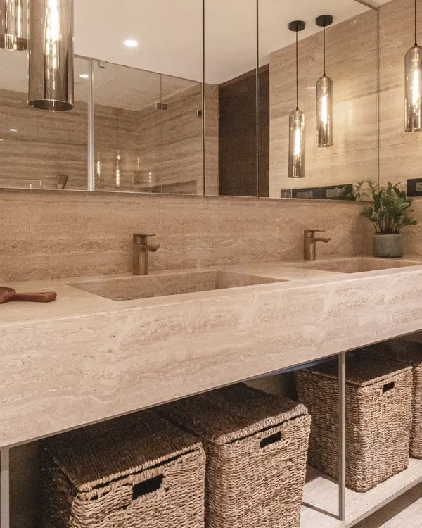 This master bathroom is a study in monochromatic luxury. A custom-built travertine vanity with integrated sinks is paired with bronze fixtures and warm pendant lighting. Wicker baskets add a natural, textural element for storage.
