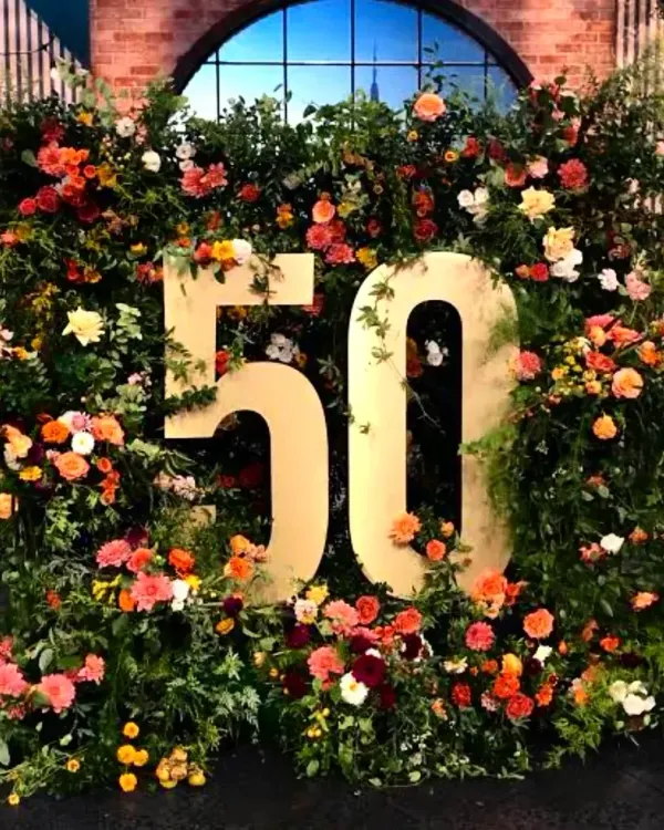 This statement photo wall was created for a 50th birthday celebration, featuring the number fifty in gold set against a lush backdrop of fresh greenery and vibrant, colorful flowers. It’s a perfect example of a grand, personalized milestone decoration.