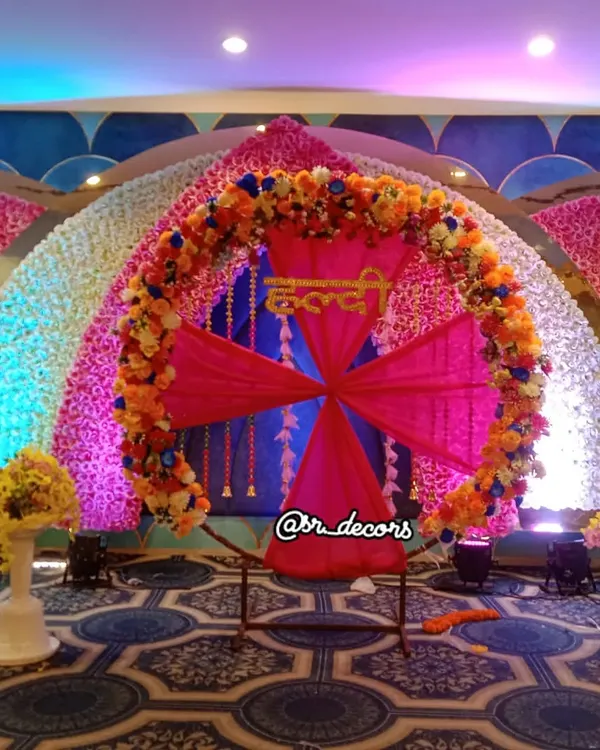 This grand Haldi stage is perfect for a banquet hall setting. The decor is centered around a large ring with 'Haldi' written in Hindi, surrounded by walls of colorful flowers.