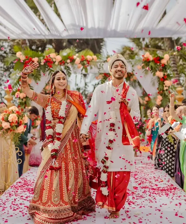 A perfect moment of joy as the newlyweds walk down an aisle showered with petals. We designed this outdoor ceremony with a flowing white fabric canopy and lush floral arrangements in red and peach, creating a romantic, garden-like atmosphere for their first steps as a married couple.