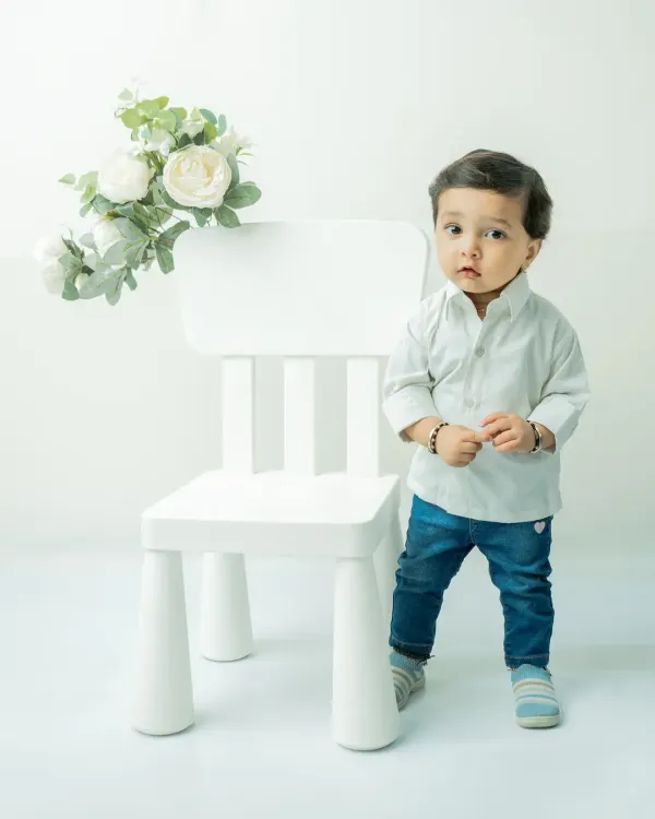A classic pre-birthday portrait of an 11-month-old boy. Standing by a simple white chair against a white background, his serious and curious expression is the main focus of the shot.