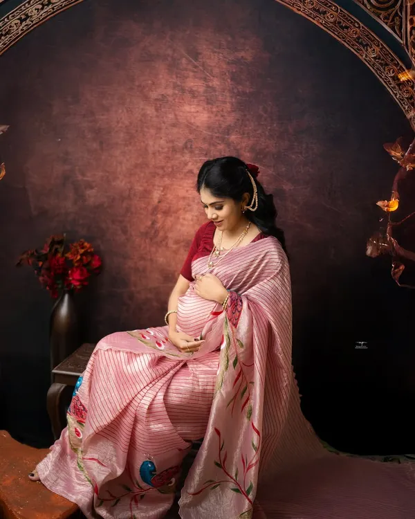 A quiet moment of connection. Seated gracefully in a beautiful pink saree, this mother-to-be gently holds her belly, creating a portrait of pure serenity and love.