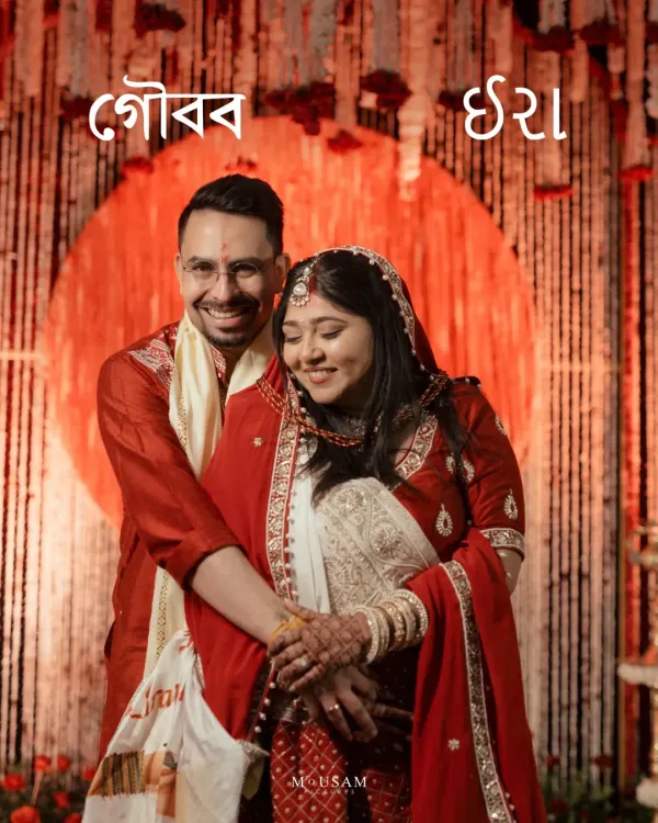 A classic portrait of a Bengali bride and groom, their names beautifully written in Bengali script. They are wrapped in a loving embrace against a backdrop of crimson and gold, a timeless frame from their special day.