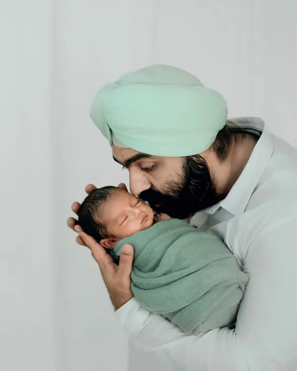 A father in a soft green turban gently kisses his swaddled newborn's head. This image celebrates the quiet, powerful bond of new fatherhood.