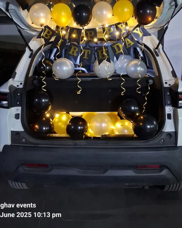 A classic car boot birthday surprise. The decoration includes a 'Happy Birthday' banner, fairy lights, and balloons in black, gold, and silver.
