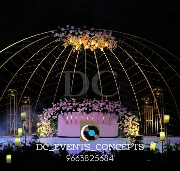 A breathtaking outdoor reception setup featuring a large, domed metal structure, soft floral arrangements, and dramatic uplighting to create an enchanting evening ambiance.